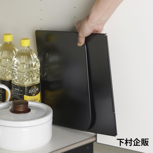 一只手正拿著一片黑色的ㄇ型折疊防汙擋油板，板面呈現霧面質感，旁邊有兩瓶食用油和一個白色鍋子。