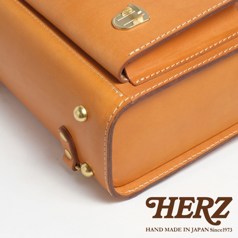 HERZ 品牌標誌，下方印有 HAND MADE IN JAPAN Since1973，整體為一只棕褐色皮革製作的橫式包款，細節處有金屬鎖扣、鉚釘和縫線。