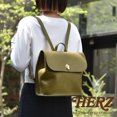 一個橄欖綠色的職人手作皮革翻蓋迷你後背包，配有金色金屬鎖扣和可調節肩帶，由一位身穿白色毛衣和黑色裙子的女性背著。