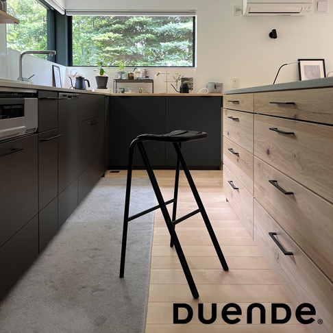 黑色金屬吧檯椅，椅面為一體成形設計，椅腳為細長金屬支架，整體造型簡約，擺放在木質地板與深色廚具之間。