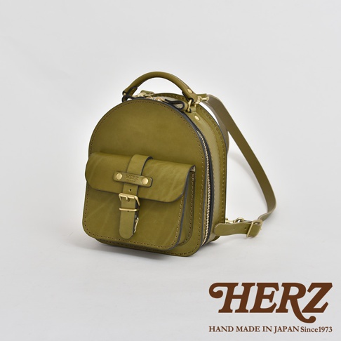 橄欖綠皮革迷你後背包，圓弧形設計，正面有一個帶有金屬釦帶的翻蓋口袋，側邊有拉鍊開合，頂部有提把，配有可調節的皮革肩帶。