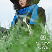 一名女子戴著黑帽，身穿深色上衣，圍著一條鮮藍色的針織圍巾，圍巾上有白色掛繩和金屬扣環。背景為綠色草地與淡藍色天空。