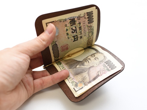 一只手拿著深棕色皮革製成的職人手作橫式對折鈔票短夾，夾內有多張日圓鈔票，鈔票上有數字「10000」及肖像。