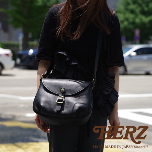 HERZ 品牌標誌與「HAND MADE IN JAPAN Since1973」字樣，中間為一名女性肩背黑色皮革馬鞍包的特寫。