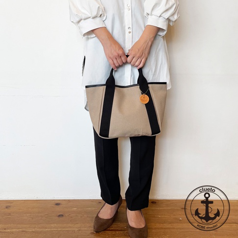 米色帆布托特包，配有黑色織帶裝飾和黑色皮革提把，包上掛有圓形皮革吊牌，風格簡約。