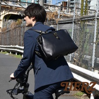 職人手作黑色皮革軟式杜勒斯包，男士著深藍色西裝騎乘自行車，包包以雙肩背方式掛在背後，配有金色金屬扣環。