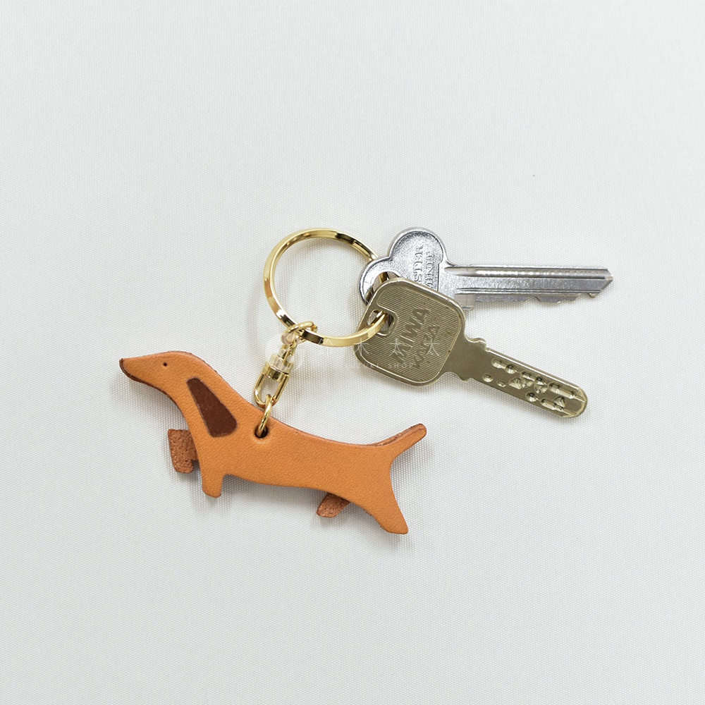 職人手作皮革臘腸狗造型鑰匙圈，淺棕色皮革主體，搭配深棕色耳朵與斑紋，附有金色鑰匙圈及兩個銀色鑰匙。