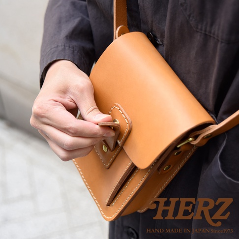 淺棕色皮革製成的職人手作散步包，包身帶有縫線細節，使用金屬釦環開合，由手部特寫展示。