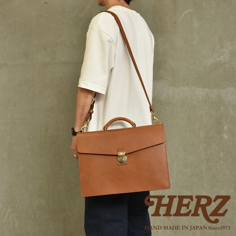 職人手作皮革Organ兩用商務公事包，採用光滑的棕色皮革製成，配有頂部提把和可拆卸肩帶，正面設有旋轉鎖扣，適合商務場合使用。