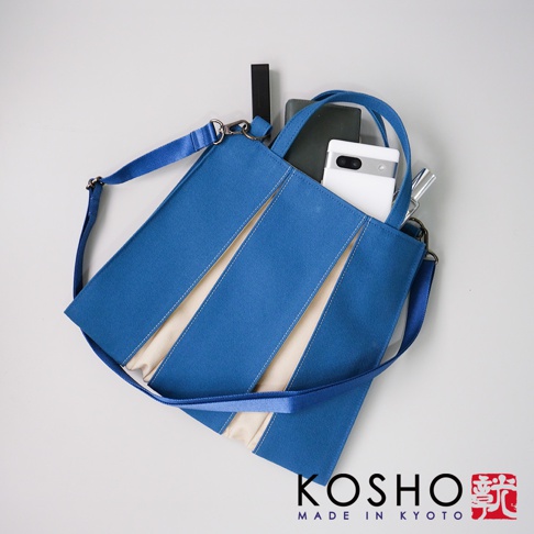 京都職人手作ougi棉帆布兩用扇形托特包，此款為藍色，包身飾有米白色斜條紋設計，使用堅韌的棉帆布材質。包包內可見手機、筆等物品，配有可拆卸的長背帶和手提把，可肩背或手提。