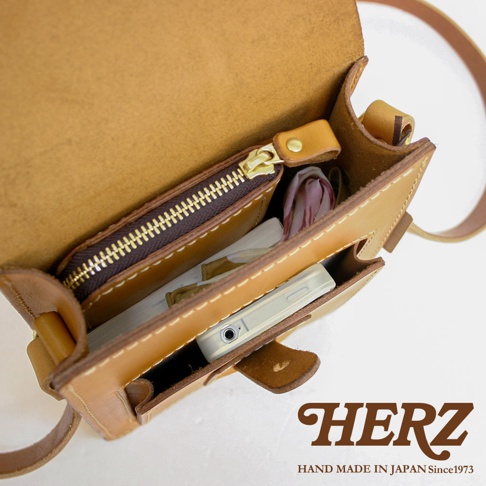 職人手作皮革休閒散步包，展示包包內部，裝有手機、卡夾和絲巾。包包為淺棕色皮革，縫線明顯，整體簡約實用。