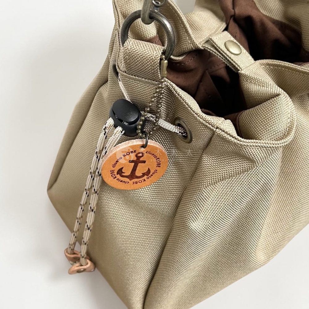 這是一款神戶 clueto 手提包的細節特寫，包身選用米色防潑水材質，袋口由抽繩設計搭配黑色調節扣固定，並掛有一枚刻有船錨圖案與 clueto KOBE 字樣的圓形皮革吊飾，整體展現簡約實用的日系風格。