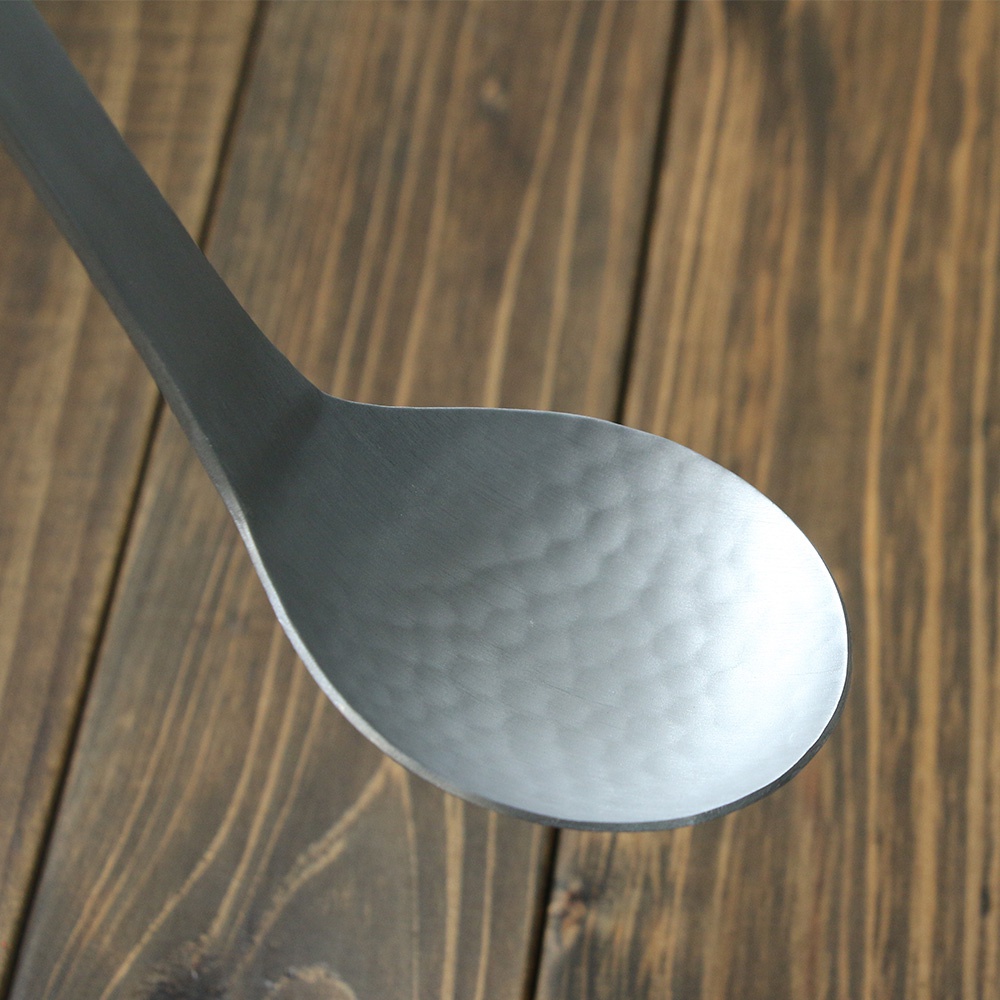 銀色鋁製湯勺，勺面帶有錘目紋理，勺柄寬闊，置於深棕色木紋背景上。