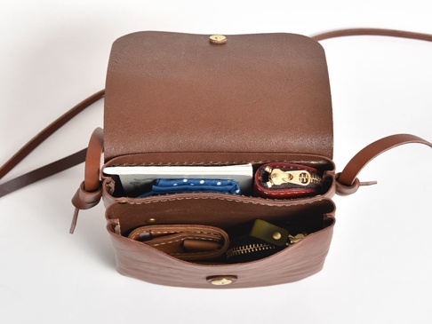 職人手作皮革直式隨行袋，深棕色，翻蓋設計，內部可見名片、藍色點點布料、紅色零錢包及錢包，材質為皮革，附有可調節背帶。