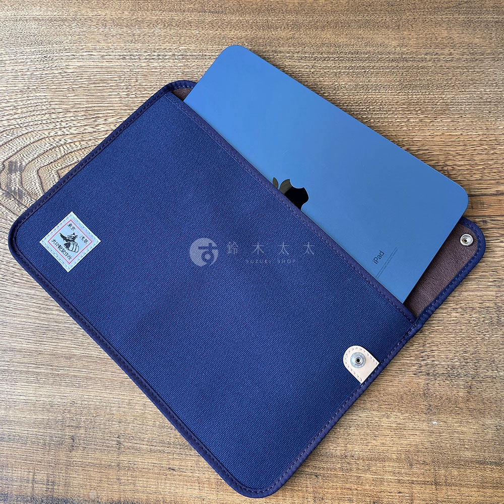 深藍色純棉帆布平板收納包，內側為棕色，包體左上角有方形品牌標籤，右下角有按扣固定。包內裝有一台藍色 iPad。