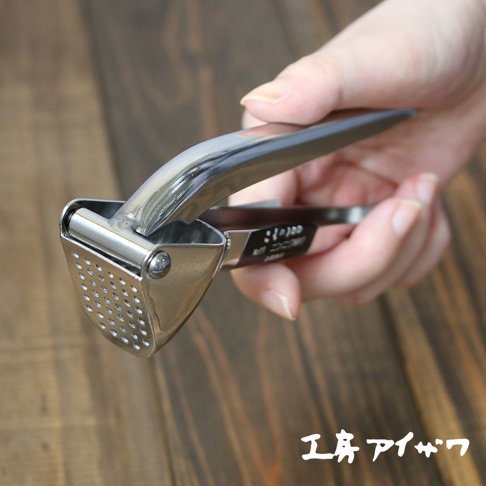 一只手正握著一個銀色的不鏽鋼壓蒜器，壓蒜器的細節清晰可見，包括其拋光的金屬表面和帶有多個小孔的壓榨腔。