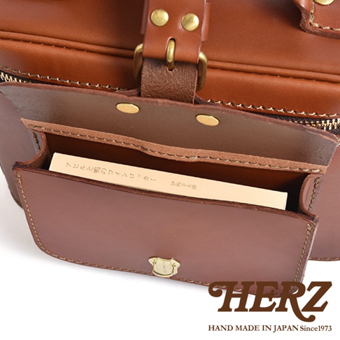HERZ 職人手作相機包，棕色皮革製作，打開的蓋口露出內部的卡片夾，上面有日文說明文字，包包下方壓印有 HERZ 品牌標誌。