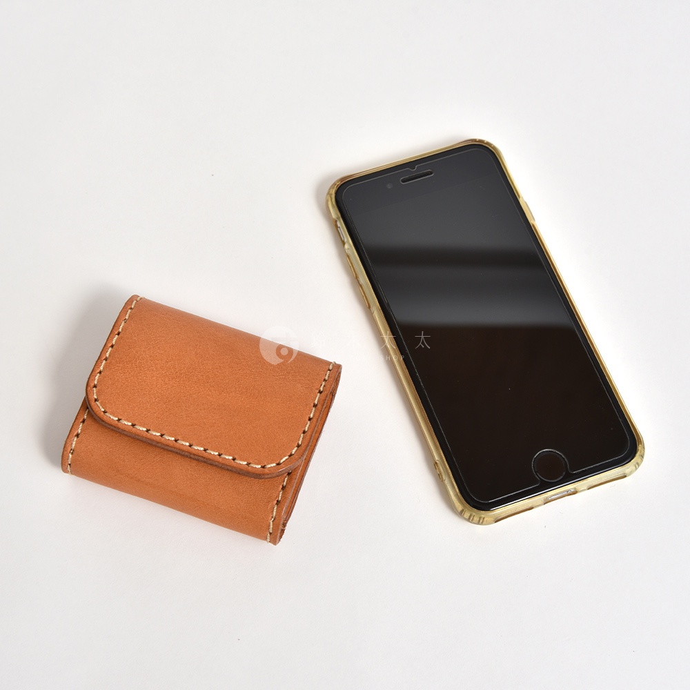 日本HERZ職人手作皮革雙折收納零錢包，選用棕色Stare皮革製作，外觀簡約並具有明顯的手工縫線細節，放置於純白背景中與智慧型手機對比呈現其輕巧尺寸。