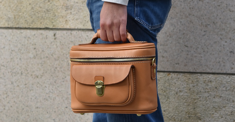 一個棕褐色皮革製的相機包，側面有一個翻蓋口袋，配有金色金屬鎖扣，頂部有一個拉鍊開口和提把。