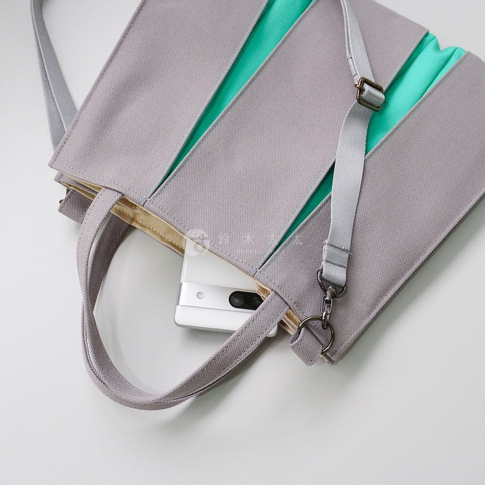 灰色棉帆布扇形托特包，內襯金色，側邊有綠色拼接設計。包包附有可調節的灰色帆布肩帶，並配有金屬扣環，可容納手機。