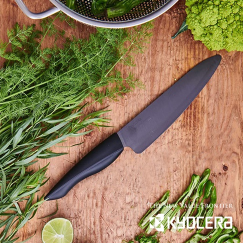 一把黑色陶瓷刀，刀刃長約 16 公分，配備黑色人體工學握把，置於木質檯面上，周圍環繞著新鮮香草、羅馬花椰菜、青檸和青椒。