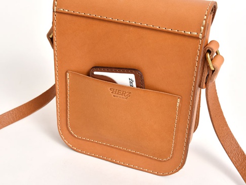 職人手作皮革翻蓋迷你單肩包，呈現天然棕色皮革質感，車縫線清晰。包款正面有一個翻蓋設計，下方有一個置物袋，袋中插有一張白色卡片。包包附有可調節的皮革背帶，搭配金屬扣環。