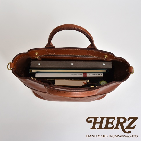 職人手作皮革大容量兩用托特包，棕色皮革材質，內部可見多本書籍、筆記本，拉鍊開合，附有提把及側邊金屬環。