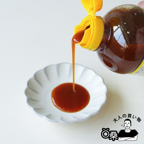 一瓶朝紫大蒜酸橘醋風味萬用調味醬的瓶口朝下，將深褐色的醬汁緩緩倒入白色的花瓣造型小碟子中，畫面右下方帶有一個男性插畫圖案與大人的購物字樣。