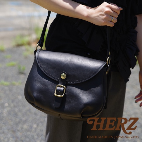 HERZ 職人手作黑色皮革馬鞍包，附有金色釦環和肩帶，包包上有品牌 LOGO 和「HAND MADE IN JAPAN Since1973」字樣。