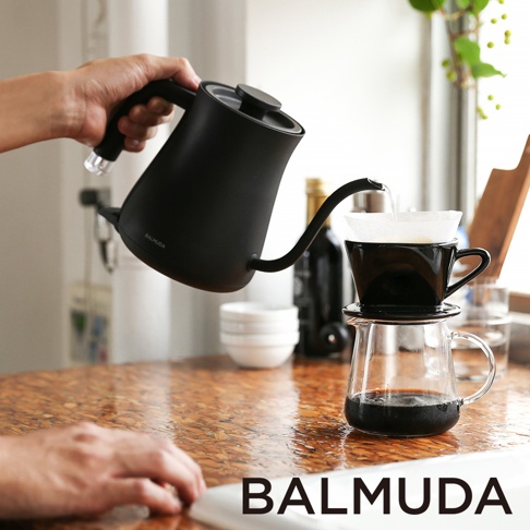 黑色啞光質感的 BALMUDA 手沖壺，壺身圓潤，配有細長彎曲的壺嘴，正傾斜著將熱水注入黑色的咖啡濾杯和玻璃分享壺中。
