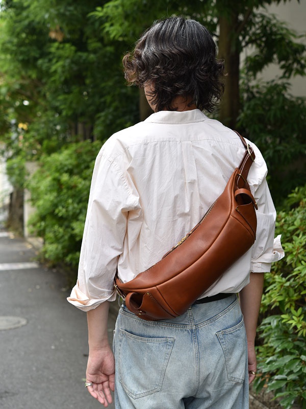 一個身穿淺色襯衫和牛仔褲的人，肩上斜背著一個品牌為 F-1 的棕色皮革大容量斜背包。