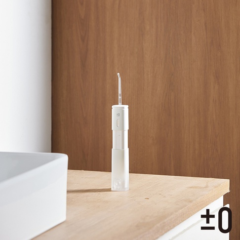 白色便攜沖牙機，機身上有 ±0 標誌與一個圓形按鈕。噴嘴為透明塑膠材質，向上延伸。產品放置於木質檯面上，背景為白色牆壁和木紋牆板。