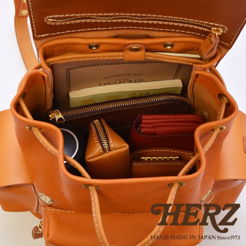 焦糖色皮革復古風旅行後背包，內部裝有筆記本、卡夾、零錢包和鑰匙包。包身由手工縫製的皮革製成，配有抽繩開合和金屬拉鍊。