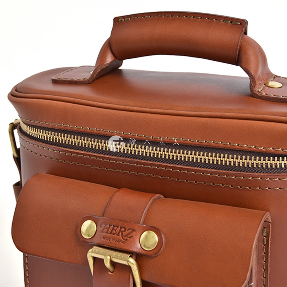 日本HERZ職人手作皮革相機包，展示了包包的拉鍊、前方口袋上的HERZ品牌標誌和Made in Japan字樣，以及包包的皮革手柄。