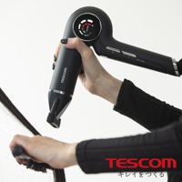 一支黑色TESCOM吹風機被一隻手握住，同時另一隻手正用梳子整理頭髮。吹風機的圓形頂部有一個顯示溫度和模式的彩色圓環，並且 TESCOM 的標誌清晰可見。