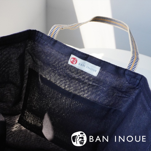 深藍色蚊帳棉織隨身購物袋，織紋細緻，配有藍黃條紋織帶提把。袋內有網狀內袋，並繡有品牌標誌。