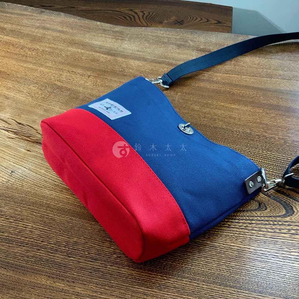 犬印純綿帆布雙色迷你側背包，採用海軍藍與紅色拼接設計，包身為帆布材質，配有可調節的深藍色織帶肩帶，設有翻蓋與金屬扣環。