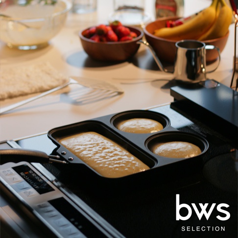 這是一款 bws SELECTION 三合一分格不沾平底鍋，正放置在廚房檯面的黑色電磁爐上，鍋內分成一個長方形與兩個圓形區域，正在烹飪麵糊。背景可見裝有草莓的木碗、放有香蕉的容器、不鏽鋼牛奶壺以及準備好的食材盆，整體呈現溫馨舒適的居家烹飪氛圍。