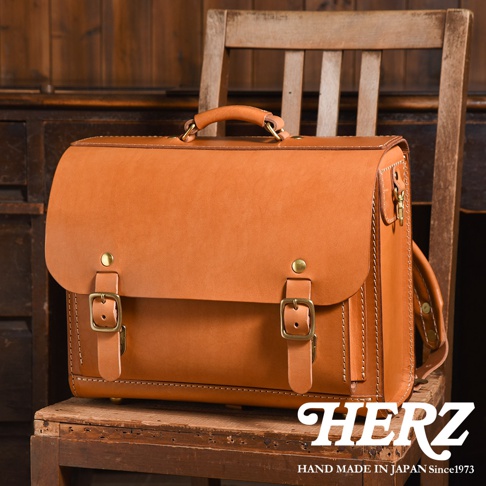 這是一個手工製作的棕褐色皮革橫式書包，有金屬扣環和手提把，放在復古木椅上。