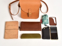 職人手作皮革翻蓋迷你單肩包，搭配棕色皮革長夾、綠色化妝包、黑色鑰匙包、手機、卡夾和一本泛黃的書籍。