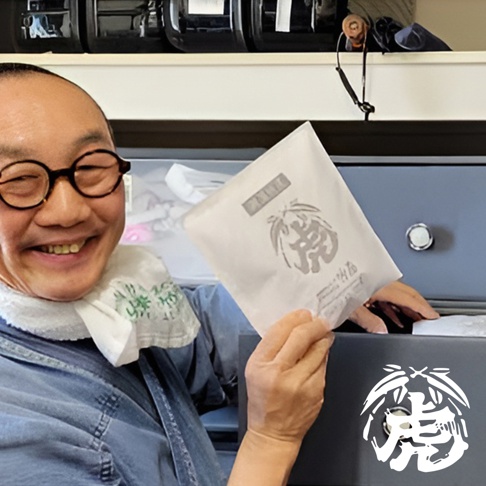 一個中型室內調濕孟宗竹炭包，白色包裝紙上印有虎形圖案和文字，由一位戴著圓形眼鏡、身穿牛仔襯衫的人士從一個灰色盒子中取出。