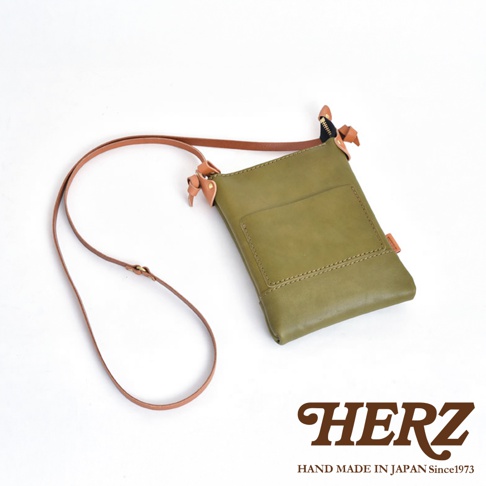 職人手作皮革直式隨身單肩包，呈現橄欖綠色，主體由皮革製成，配有可調式皮質背帶，包身前方有一個方形置物袋。