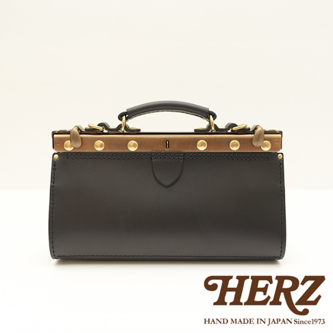 黑色皮革復古口金手提包，配有金屬鎖扣、黃銅色鑲邊和提把。包身有縫線裝飾，正面中央有一個盾牌形狀的皮革飾片。