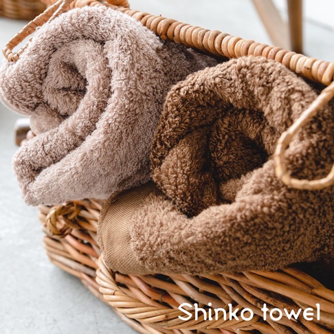 藤編籃中疊放著兩條 MAISON BLANC 輕奢柔膚匹馬棉浴巾，一條為淺褐色，另一條為深褐色，質地蓬鬆柔軟。