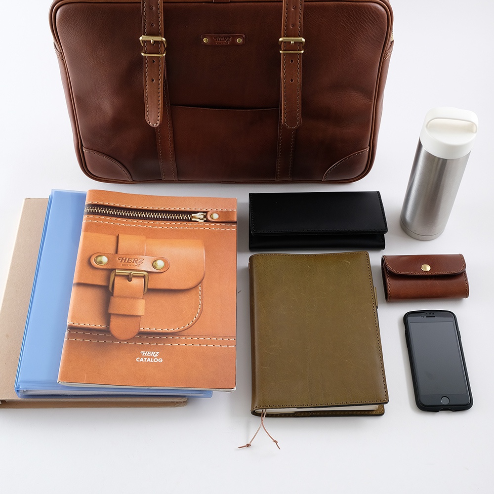 一件深棕色皮革公事包，配有雙提把與金屬扣。周圍擺放著一本棕色皮革筆記本、一本綠色筆記本、一個黑色皮夾、一個棕色零錢包、一支手機、一個銀色保溫瓶，以及幾本文件夾。