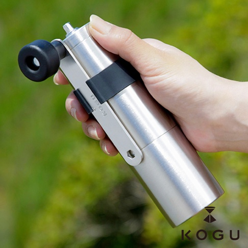 手持一個銀色圓柱形日製18-8不鏽鋼手搖咖啡磨豆器，帶有黑色旋鈕和黑色束帶，背景為模糊的綠色植物。