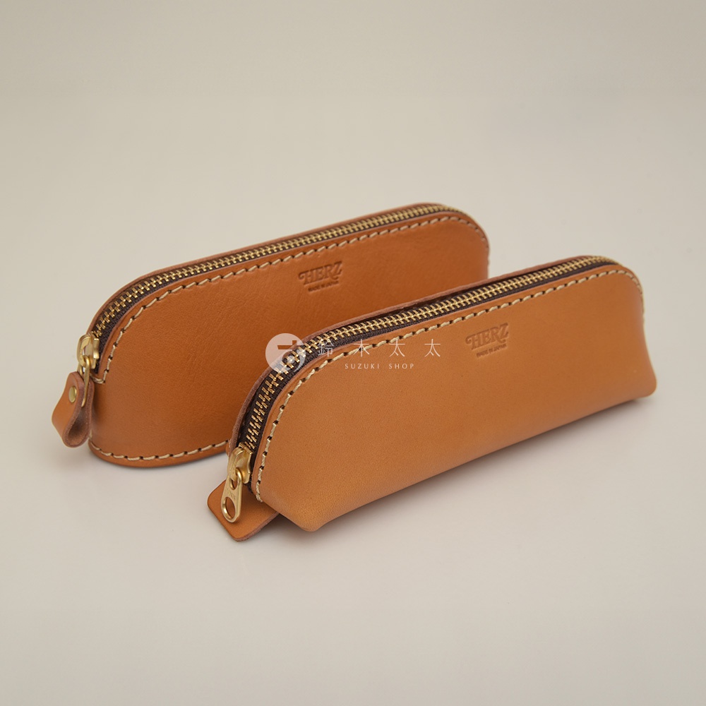 兩款棕色皮革製成的職人手作三角弧形收納筆袋，配有金色拉鍊，其中一款筆袋上有「HERZ」品牌標誌。
