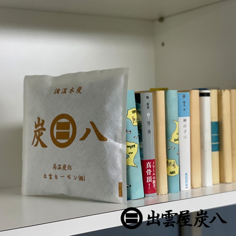 一個白色方形的出雲屋炭八調濕木炭包放置在書架上，左側為印有品牌名稱與說明文字的炭包，右側整齊排列著數本精裝書籍，整體呈現簡約的居家收納風格。