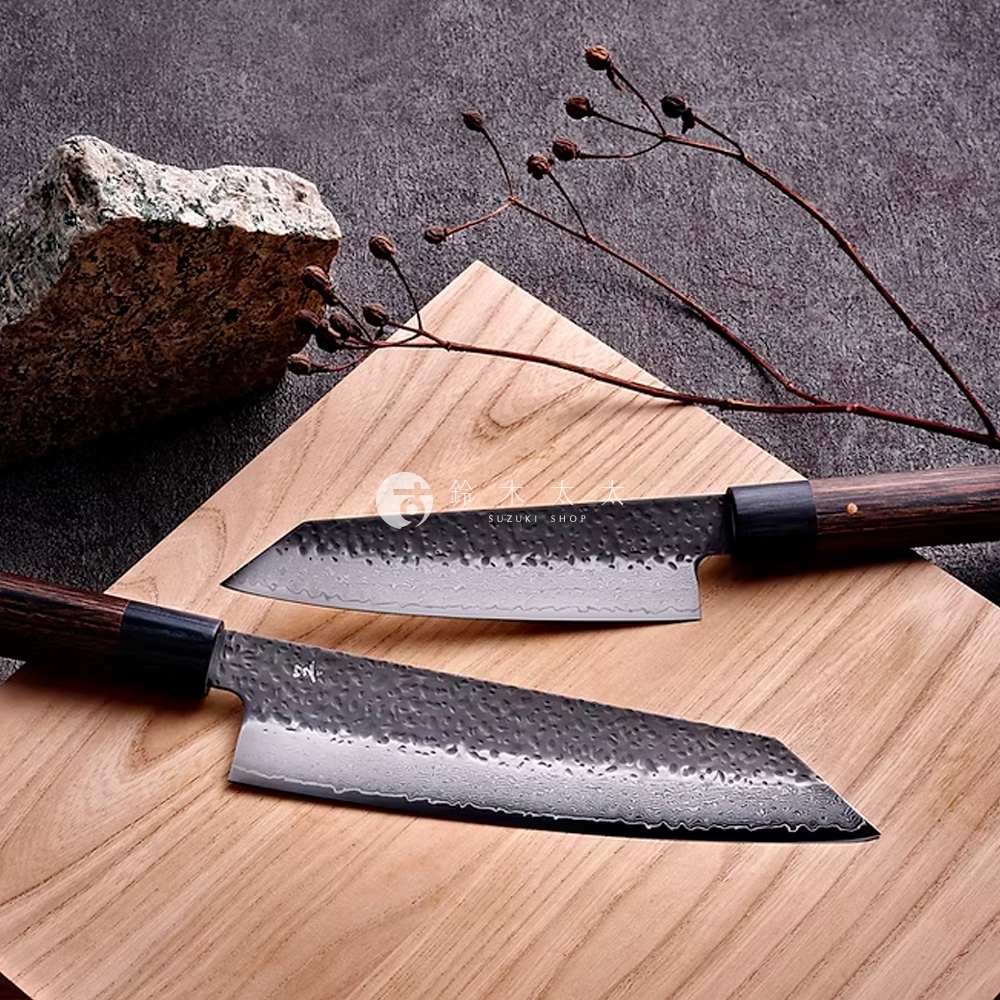 兩把「玄 / Bunka 文化刀」，刀刃呈銀白色，帶有錘紋與 Damascus 紋路，刀柄為深棕色木質，搭配黑色刀頸，擺放在淺木色砧板上，旁有枯枝與岩石裝飾。