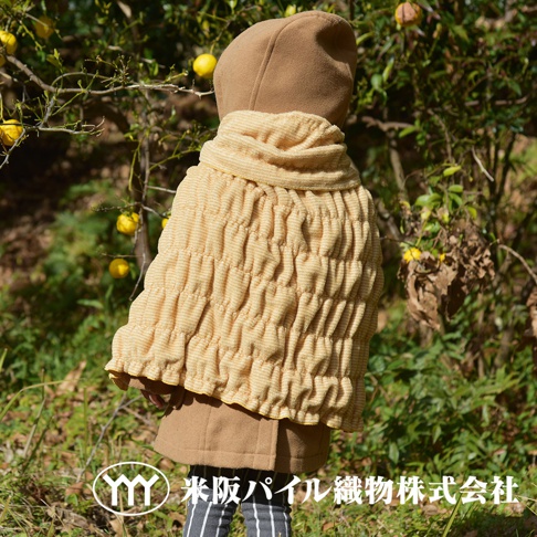 身穿駝色連帽外套，披著一件淺黃色細絨波浪紋樣的室內外兩用毯，毯子上帶有細條紋的織紋，營造出蓬鬆的褶皺感。